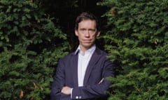 Rory Stewart wearing a navy suit and white shirt and standing in a gap in a conifer hedge