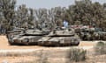 Israeli tanks form a circle on a sandy, dusty road, near to some trees. One tank in the foreground has an Israeli flag