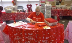 A woman in Valentine's-themed clothes sits at a table and stamps cards surrounded by Valentine's Day decorations and crates full of letters.