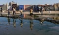 Children walk single file between patches of water, with temporary shelters in the background.