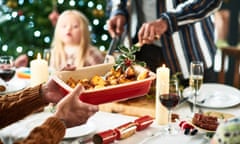 Man holding a serving dish with roast potatoes and a sprig of holly at a Christmas dinner.