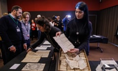 A female conservator holds up an old letter above a box containing a bundle of similar letters. Around her on tables are other documents from the collection on display