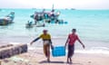 Two Arab men carry a crate of fish up a beach, as others wade into the sea to unload the catch from a fishing boat