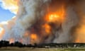 A partly sunny sky is almost completely blocked by plumes of smoke rising amid fire from a low hillside, with homes in the foreground.