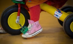 A child's foot in a pink sneaker dangles below the pedal of a yellow trike on a wooden floor