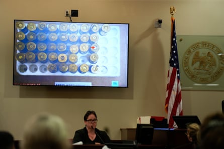 Poppell sits under a tv screen in a courtroom, next to US flag