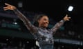 Biles stands, grinning, with her arms outstretched after her routine. She wears a shiny, metallic-effect blue, navy and black leotard. The top half of it has stars across the front. There are crystals across the top half of her leotard and on the arms