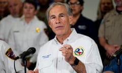 Middle-aged white man in white shirt with Texas emblem on left breast gestures and speaks into microphone with rows of people in casual uniform standing behind him.