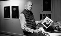 An elderly white man with a sombre face stands in a studio holding a black-and-white photograph with other prints on the wall behind him