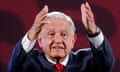 Older white man with white hair in dark suit speaks on well-lit stage, gesturing with both hands raised.