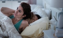 A woman sitting up in bed rubbing her neck, as her partner lies sleeping beside her