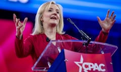 Blond middle-aged woman wearing pink at lectern raises both hands.