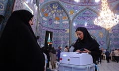 A woman watches as another woman uses a voting machine in a large decorated hall with other voters in the background