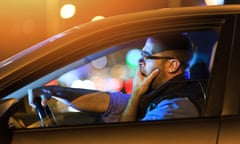Man sitting in car and yawning by night<br>Side view of young man with eyeglasses sitting inside of his car and yawning. One hand on mouth. Bright lights at background.