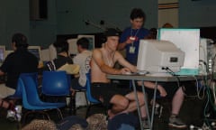 A group of young men sitting at tables in a school hall with assorted computers on the desks and clothes and sleeping bags on the floor