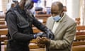A police officer wearing gloves and a visor attaches handcuffs to the wrists of Paul Rusesabagina, who is wearing a beige suit and blue surgical mask