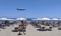 A passenger plane approaches Larnaca airport, Cyprus, as a woman walks past sunbeds on a busy beach.