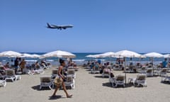 A passenger plane approaches Larnaca airport, Cyprus, as a woman walks past sunbeds on a busy beach.