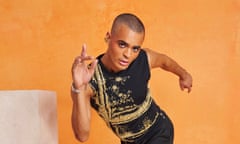 Layton Williams posing on one leg and leaning forwards, as if he is dancing, wearing a black vest embellished with gold, a gold earring, black trousers and boots, and photographed in an orange space with white marbled blocks