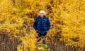 An older man with bright white hair and moustache in a blue-and-black checked shirt stands with his hands in his pockets amid stunningly bright yellow trees.