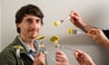 A man smiling against a beige background, while hands hold forks with ravioli speared on them.