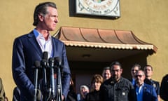 man wearing blue suit stands with hands clasped in front of five microphones as people stand behind him