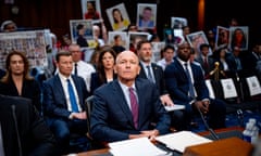 man wearing suit and tie sits at table in front of rows of people holding up portrait photos