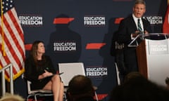 Sarah Huckabee Sanders sits n stage next to a flag with a backdrop printed with the shape of Tennessee and the words "Education Reform" while Bill Lee speaks.