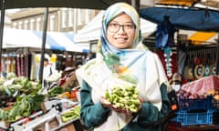 Nadia Mohd-Radzman, with glasses and in a headscarf and cotton jacket, stands in front of vegetable market stalls holding a bunch of broad beans and grinning