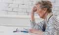 An older woman sits at a desk with a calculator looking worried.