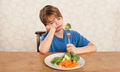 A boy sitting at a table with a plate full of vegetables frowning at a piece of broccoli on a fork