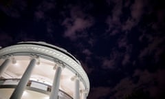 In the bottom left corner, the camera looks up into a brightly lit, two-story, white rotunda. The rest of the frame is a nighttime sky with a few sparse clouds.