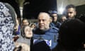 A middle-aged Palestinian man with a bald head, light white bread, blue flak jacket, looking stunned and standing in a crowd of people, with a young woman, her hair in a scarf, chin raised and crying beside him.