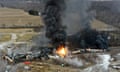 An aerial view of piled-up train cars, with a fire raging in the middle of them and sending up plumes of black smoke, above farmland and woods.