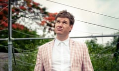 Damien Power is standing under a hills hoist looking at the sky. He is wearing a white shirt and a pink and white checked jacket