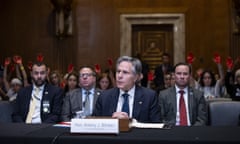 Man sits in committee room with people behind him holding up their palms covered in red paint