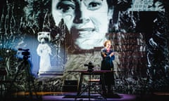 Riana Duce as Mina Harker in a trenchcoat, in front of a screen with a huge projection of her face.