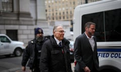 man in a black puffer jacket walks with a police officer behind him and a man in a suit next to him