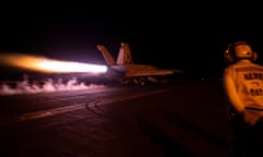 A fighter jet takes off from an aircraft carrier at night as a crew member looks on.