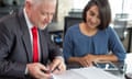 Content coach reviewing notes of female company newcomer. Middle aged man in tie and young Indian woman in casual wear thumbing notebook in coworking space.