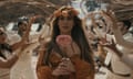 woman in sparkly burnt orange dress holding a dahlia and wearing a flower crown with dancers in white behind her