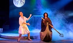Toby Park in a toga, gesturing and laughing, next to Aitor Basauri in a cloak, standing on a boat with an oar, with mist on the stage and a picture of the moon in The Frogs