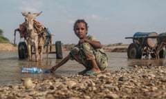 A girl collects water in a plastic bottle from a shallow river next to a donkey pulling a cart