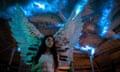 A girl wearing a white dress and angel's wings stands in a room with supernatural light coming through cracks in the ceiling.