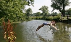 Man and a dog jumping into a river. He is diving head-first and wears blue patterned shorts; the dog is a golden labrador. A rowing boat and people on the bank can be seen in the background as the river curves, with trees and a field to the side.