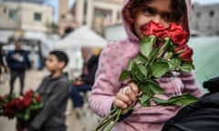 A Palestinian child in a pink jumper smells a bunch of red roses bought from a boy selling them in the background