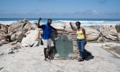 A young African couple pose on a rocky coastline in South Africa by a plaque marking Africa's southernmost point