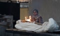 A young woman stands in a large building with hands raised in prayer as a shrouded corpse lies at the door of a furnace, with flames seen from a burning firebrand