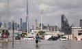 A person appearing to hold their shoes in one hand walks knee deep in water along a city street, with high rises in the background.