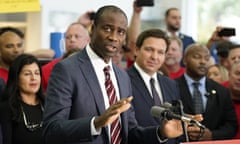 A middle-aged Black man with closely trimmed black hair, wearing a dark suit, white shirt, and dark red tie with white stripes, stands at a lectern, speaking and gesturing, with Ron DeSantis beside and behind him, among a large group of mostly smiling people.